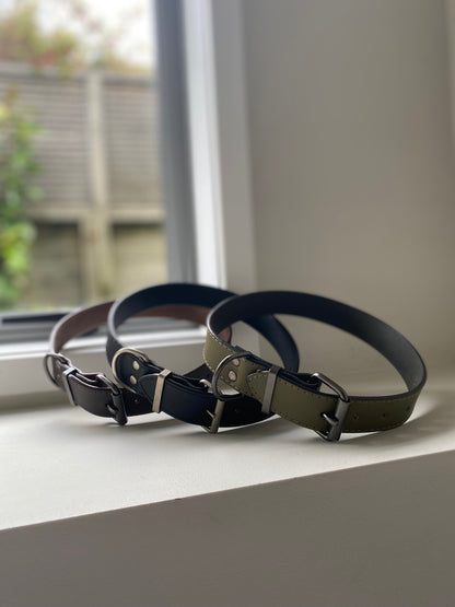 Three leather dog collars on a windowsill with a blurred outdoor background. One of them is brown, one of them is brown and the other is a khaki green.