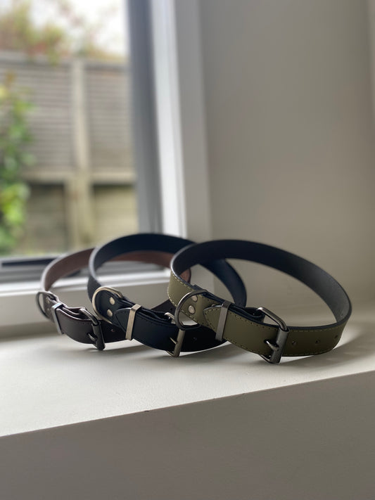 Three leather dog collars on a windowsill with a blurred outdoor background. One of them is brown, one of them is brown and the other is a khaki green.