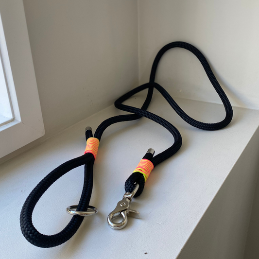 Black rope dog leashes with metal clips on a white surface. It has got neon yellow and pink paracord wrapped around both areas where the leash joins for a pop of colour.
