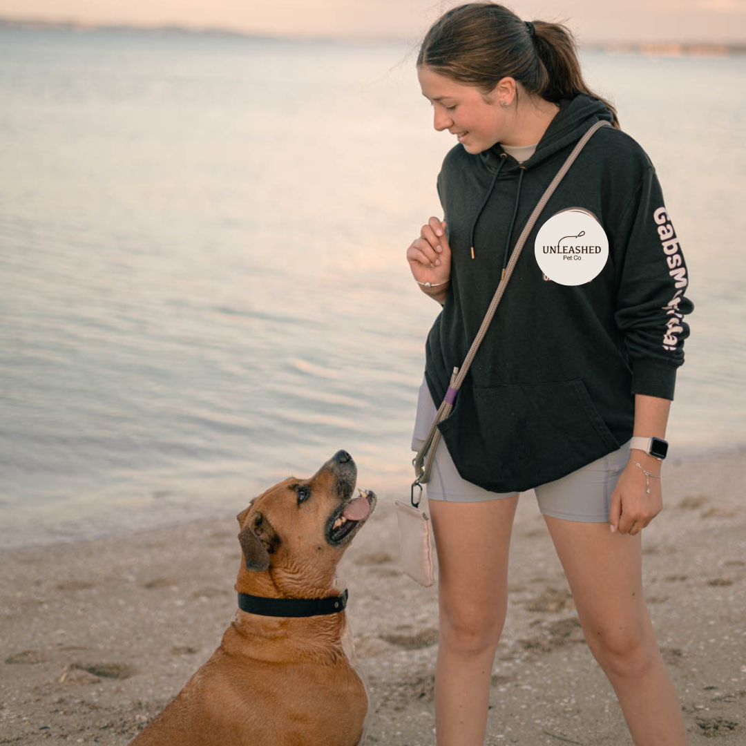 Woman in a black hoodie with 'UNLEASHED' logo standing on a beach training her dog, the leash is wrapped across her body and the dog is sitting next to her. 